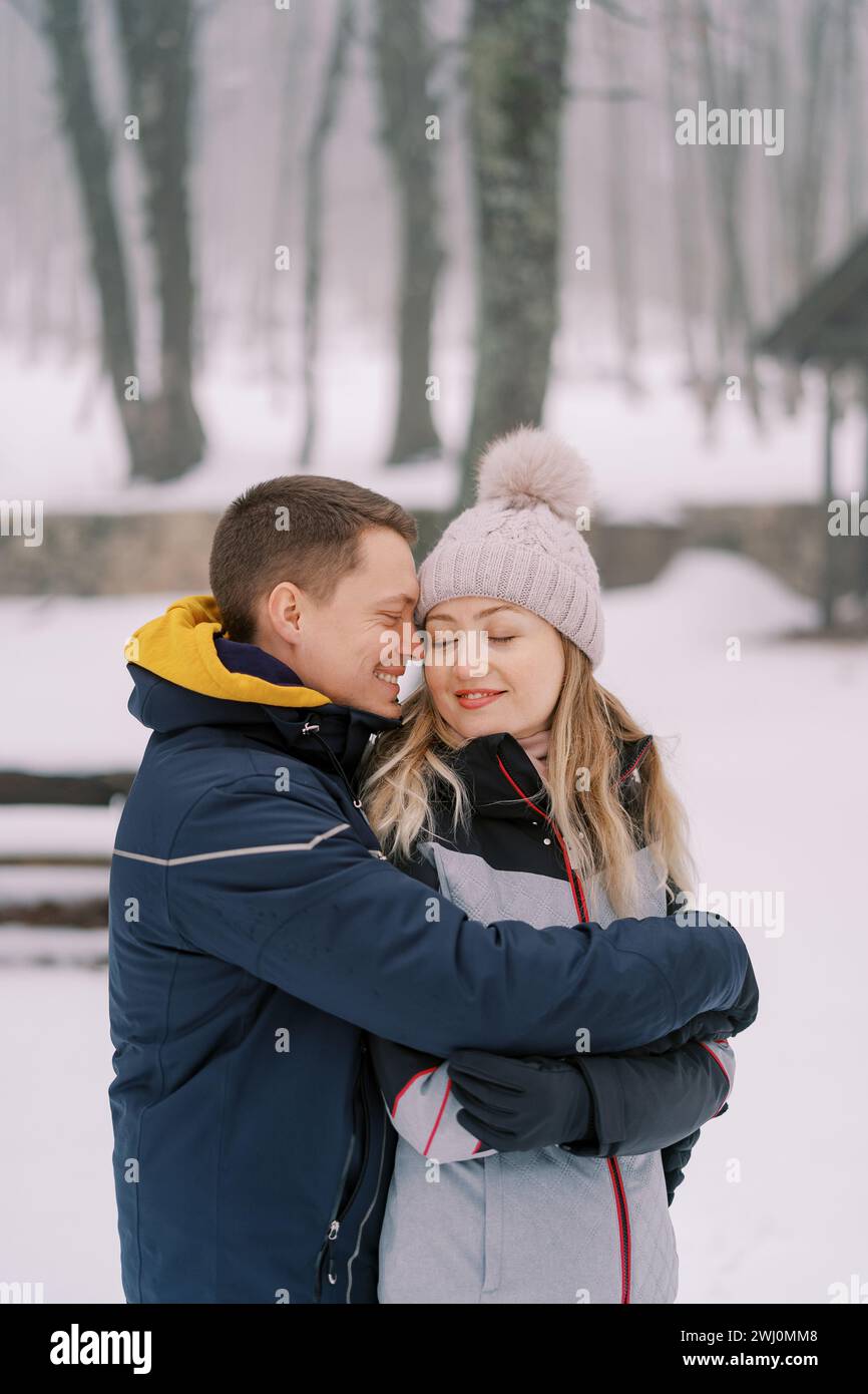 Petit ami souriant et petite amie serrant tout en se tenant dans un parc enneigé Banque D'Images