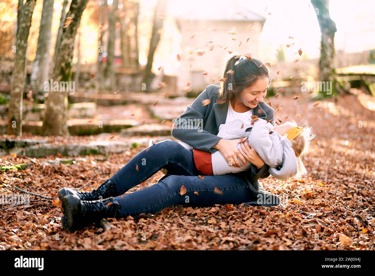maman souriante embrassant une petite fille sur ses genoux tout en étant assise dans le parc d'automne sous les feuilles tombantes Banque D'Images