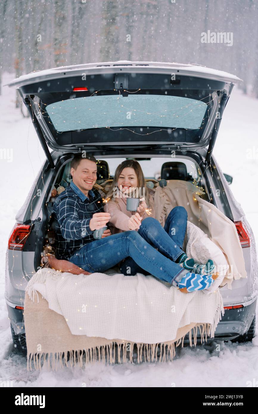 Souriant petit ami et petite amie sont assis dans le coffre d'une voiture sous la neige avec des étincelles et des tasses de café Banque D'Images