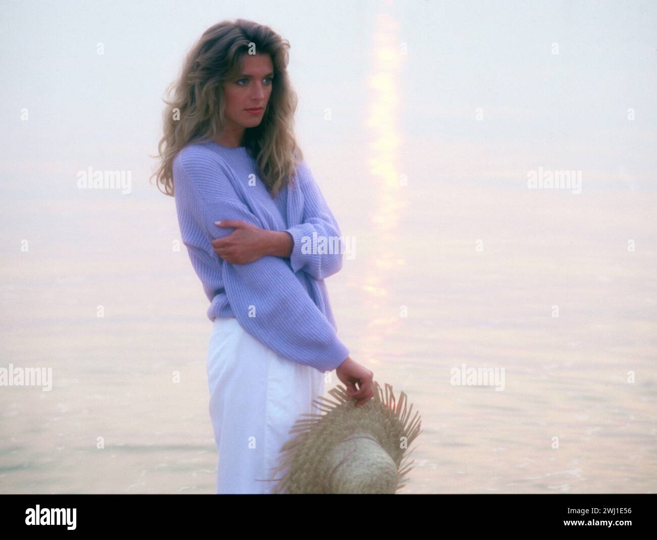 Jeune femme aux cheveux blonds debout près de l'eau au coucher du soleil. Banque D'Images