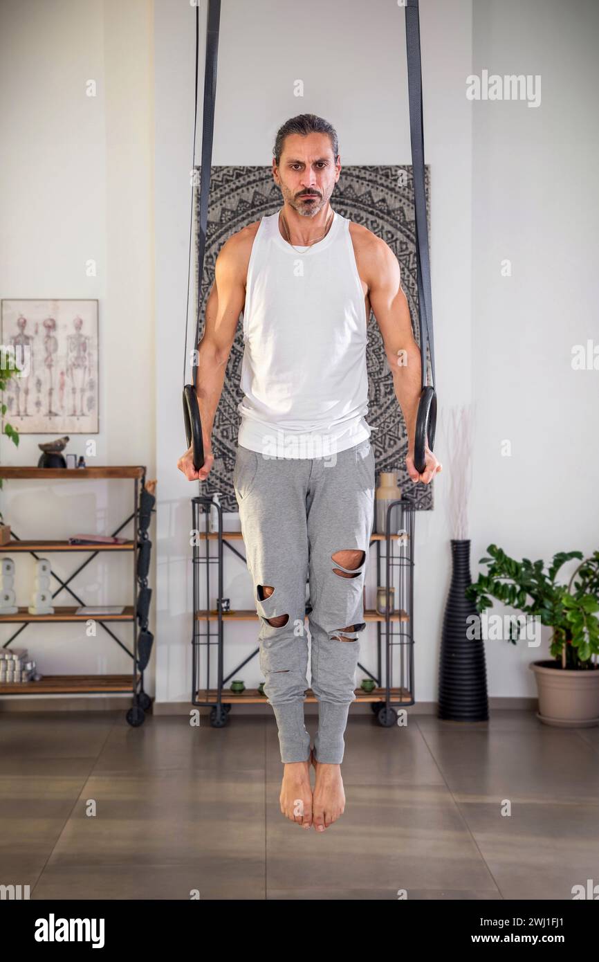 Corps entier d'un homme adulte confiant pieds nus avec la barbe regardant la caméra tout en plongeant sur des anneaux de gymnastique dans la salle de gym et en faisant de l'exercice à la lumière du jour Banque D'Images