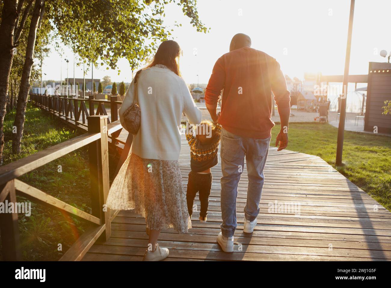 Vue arrière de la famille avec petit enfant marchant dans le parc, tenant les mains de bébé au coucher du soleil, dans les rayons du soleil couchant. Maman, papa et fils au milieu. Banque D'Images