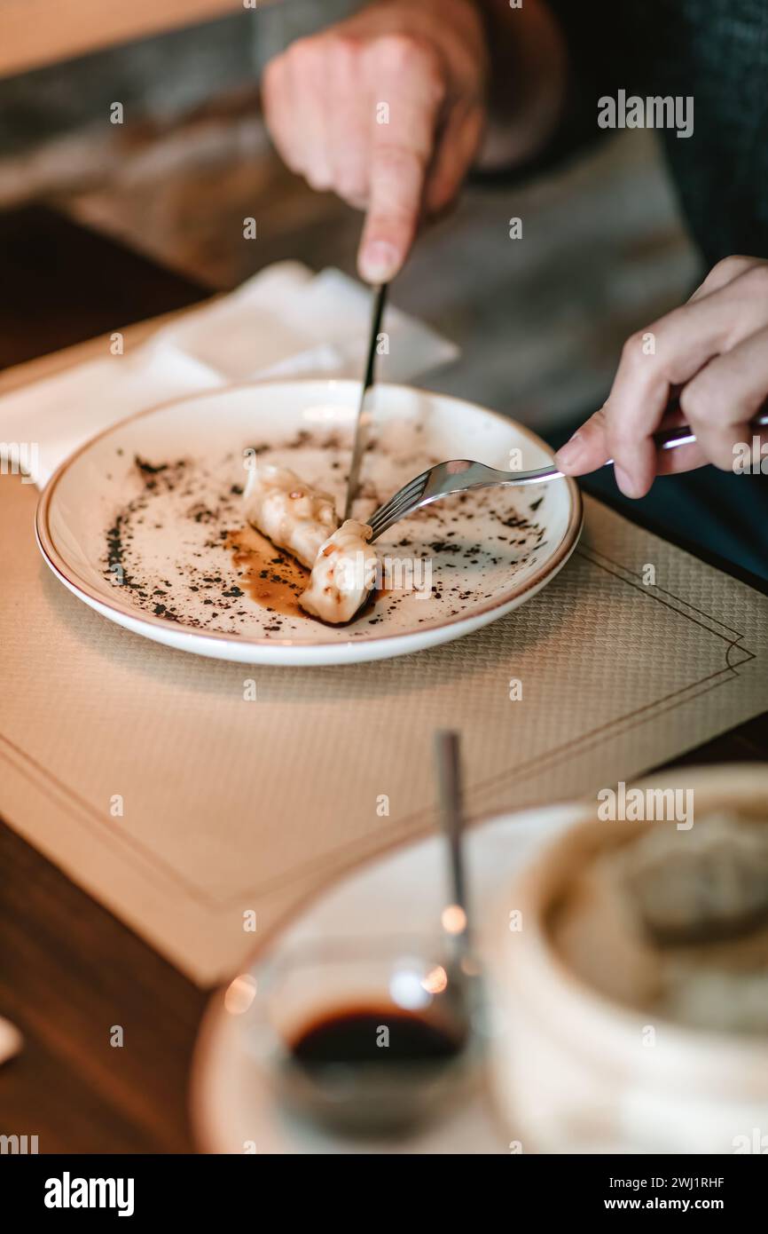 Gros plan des mains avec un couteau et une fourchette coupant un gyoza sur une assiette design, posé sur une nappe en papier et une table en bois Banque D'Images