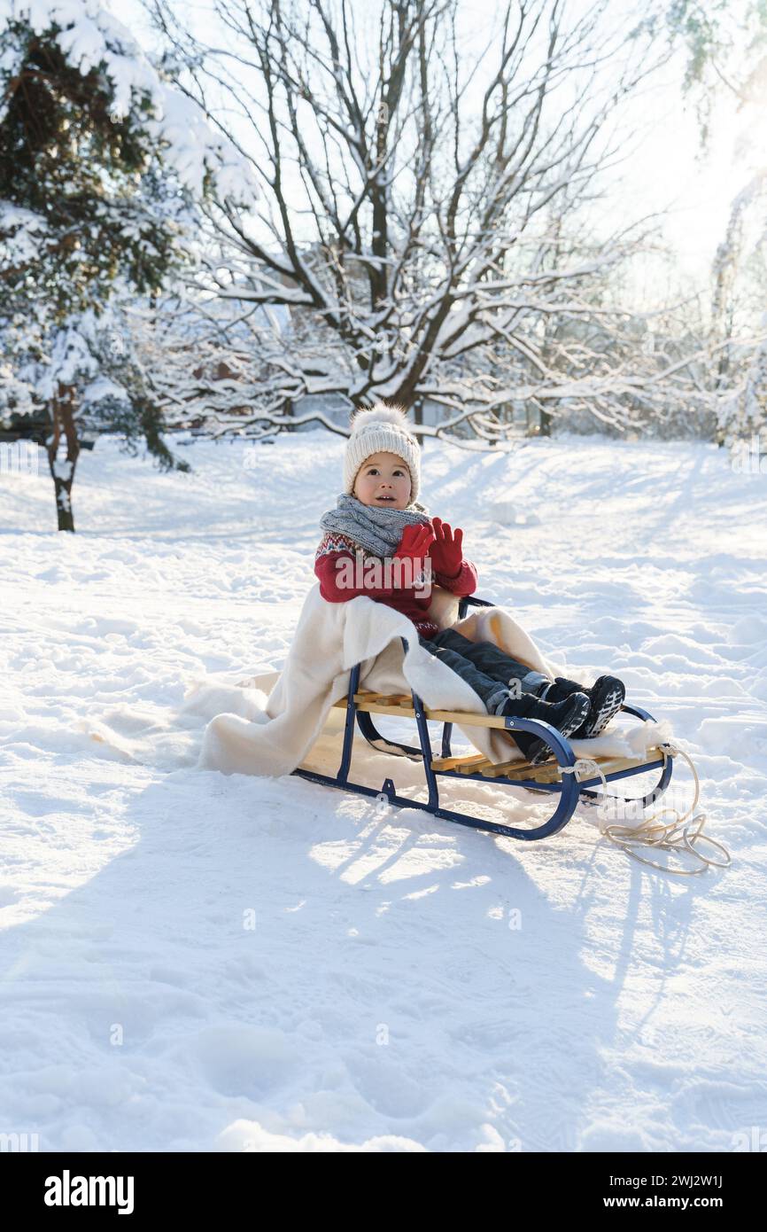 Garçon tout-petit assis sur le traîneau dans un parc enneigé de la ville pendant la journée ensoleillée d'hiver Banque D'Images