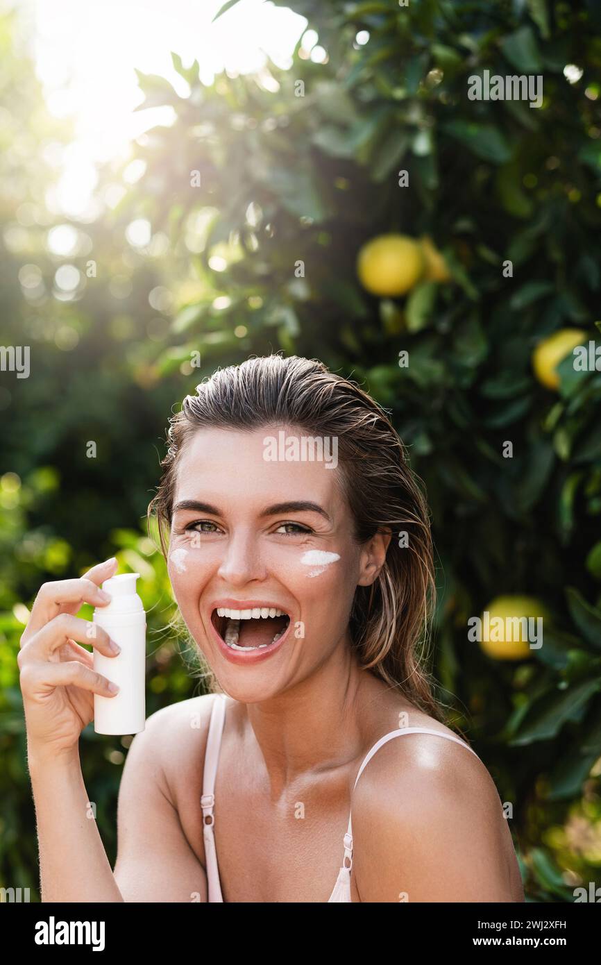 Femme joyeuse et belle appliquant une crème hydratante ou un écran solaire sur sa peau du visage Banque D'Images