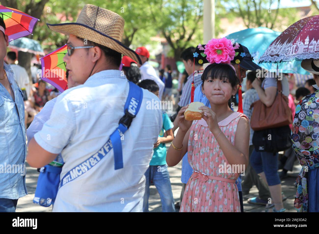 Nourrir la Chine. Une adolescente dans une rue de Pékin, une fille chinoise avec une fleur de décoration sur le dessus de sa tête mange un chignon avec de la crème à côté de papa. Banque D'Images