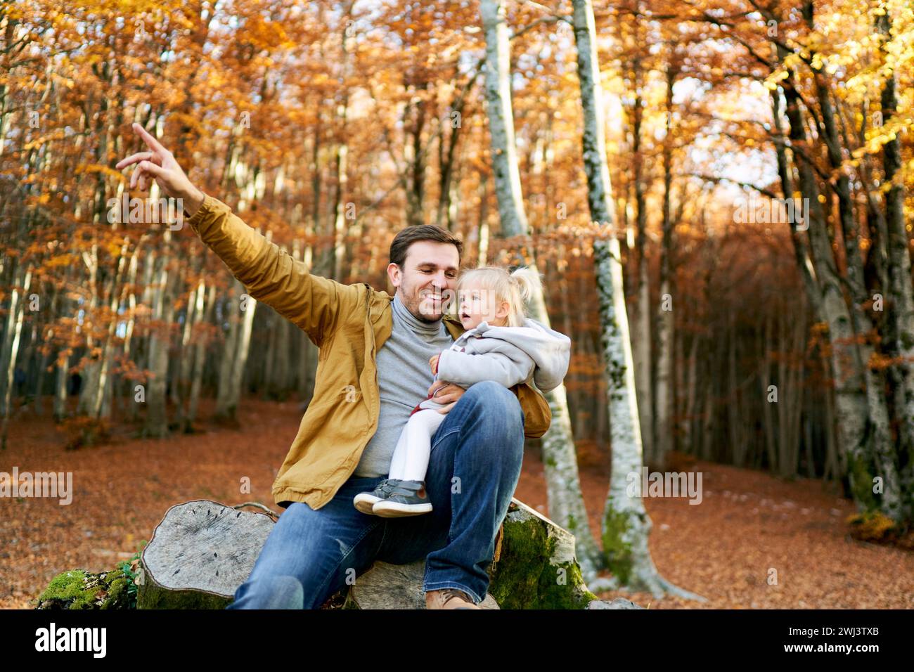 Papa souriant avec une petite fille à genoux assis sur une souche et pointe son doigt vers un arbre Banque D'Images