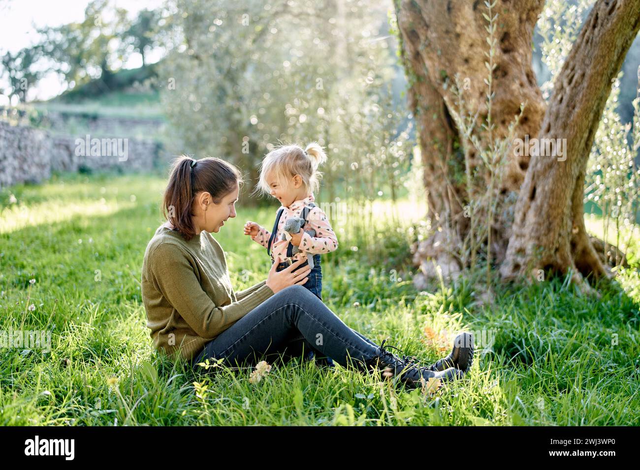 Petite fille souriante se tient près de sa mère assise sur l'herbe verte près de l'arbre Banque D'Images