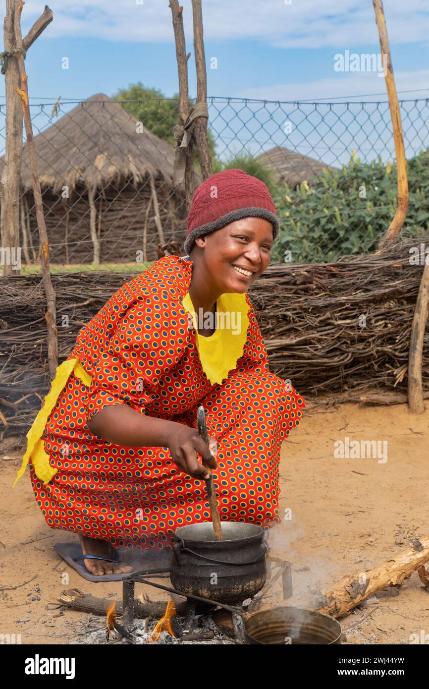 village femme africaine cuisinant dans la cuisine extérieure, cabanes avec toit de chaume en arrière-plan Banque D'Images