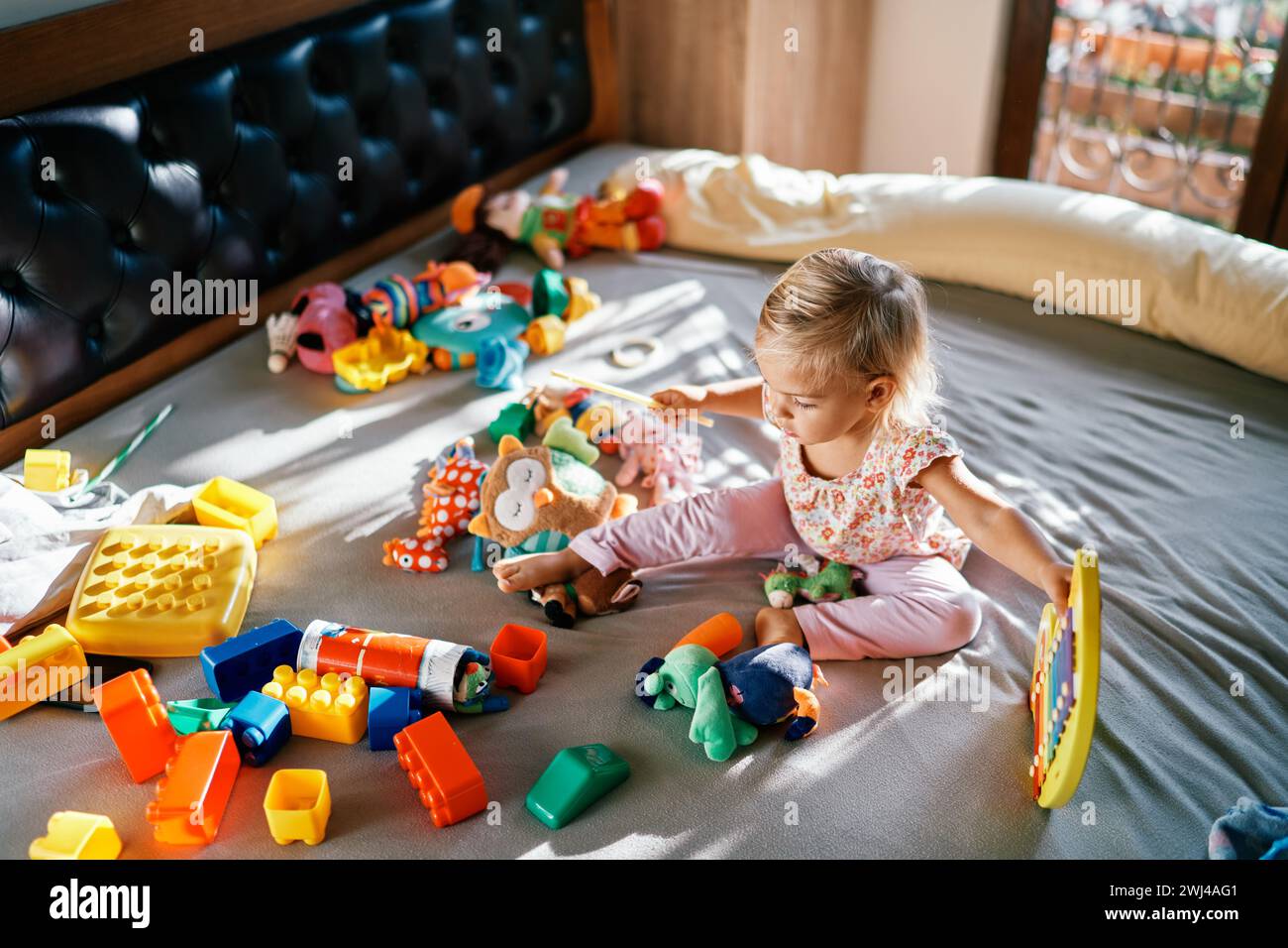 Petite fille assise avec un xylophone et une baguette dans ses mains sur le lit entourée de peluches Banque D'Images