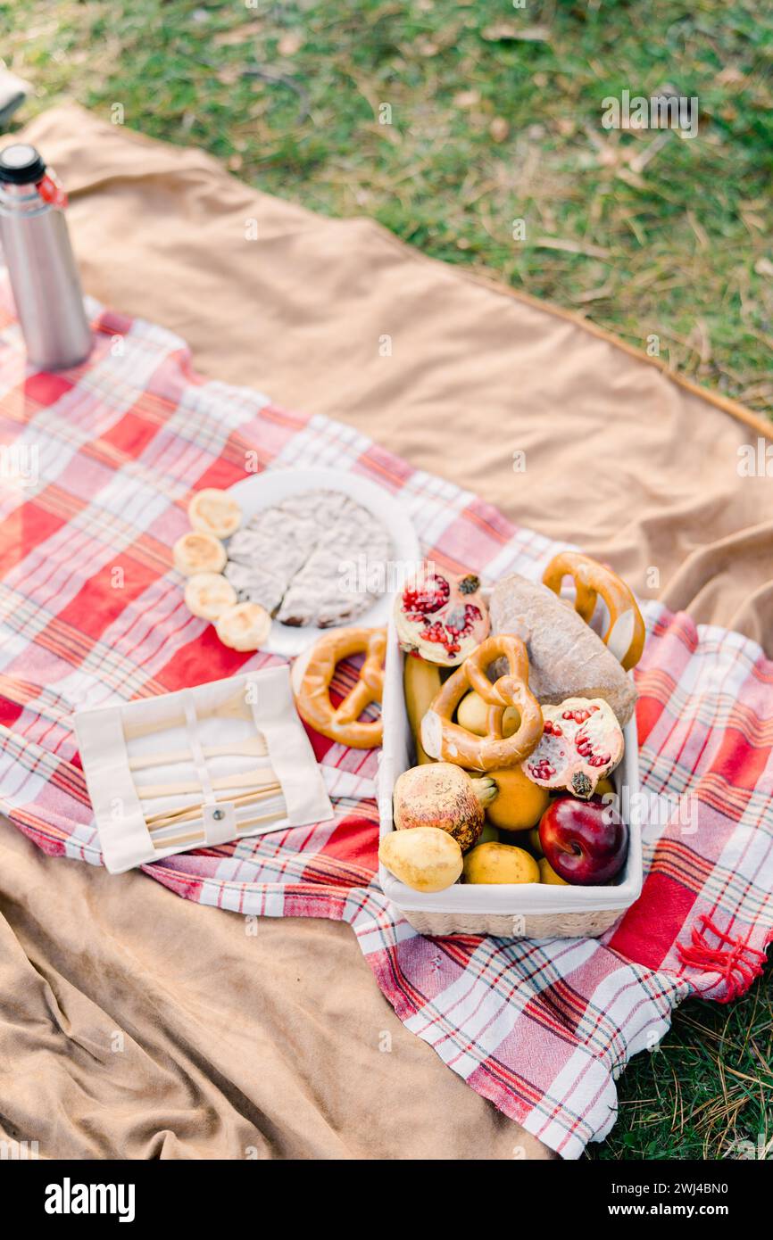 Panier de petits pains et de fruits se tient sur une couverture à côté de couverts et un thermos Banque D'Images