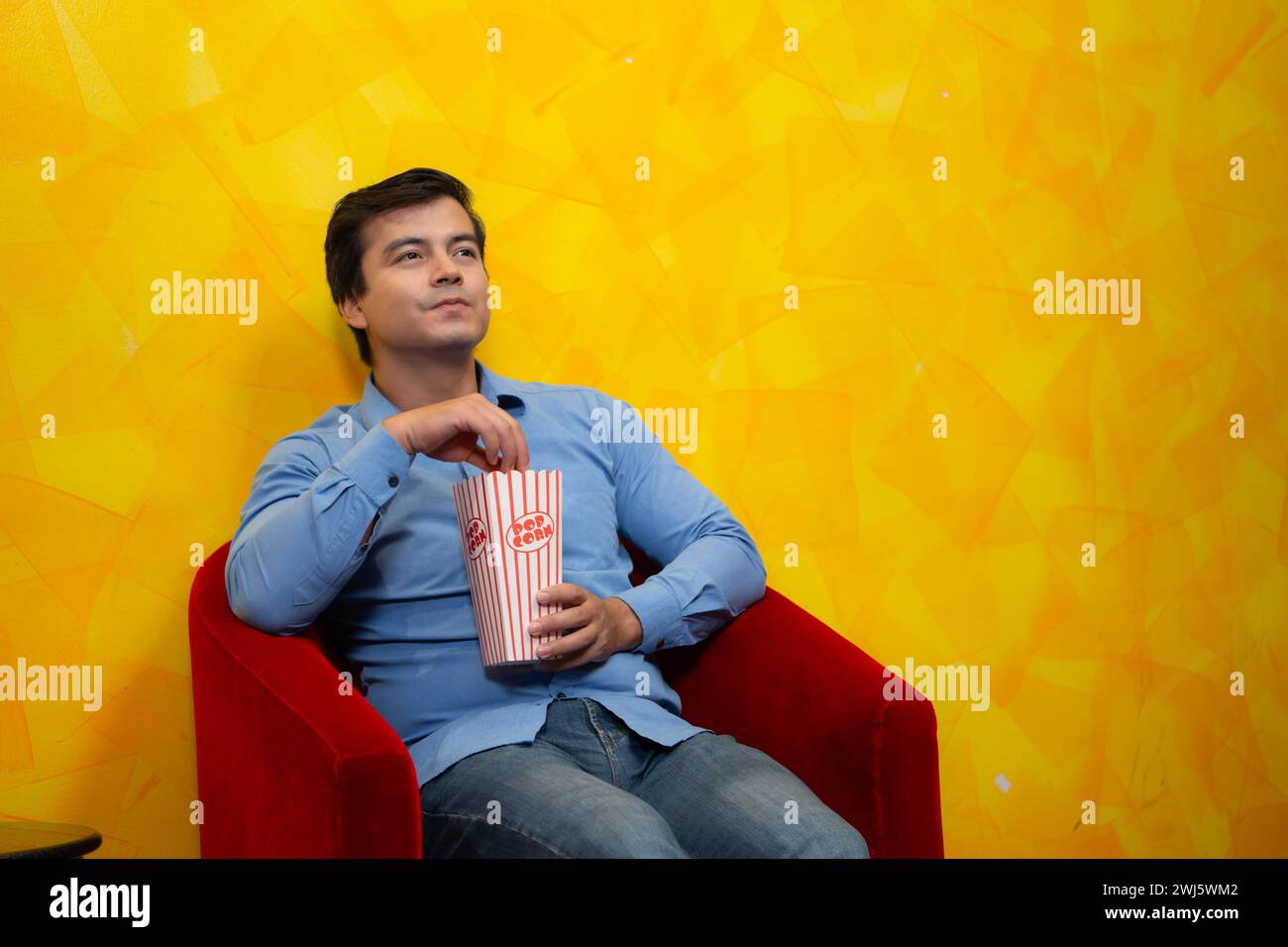 Jeune homme mangeant du pop-corn assis dans un fauteuil rouge sur un fond jaune attendant de regarder un film. Banque D'Images