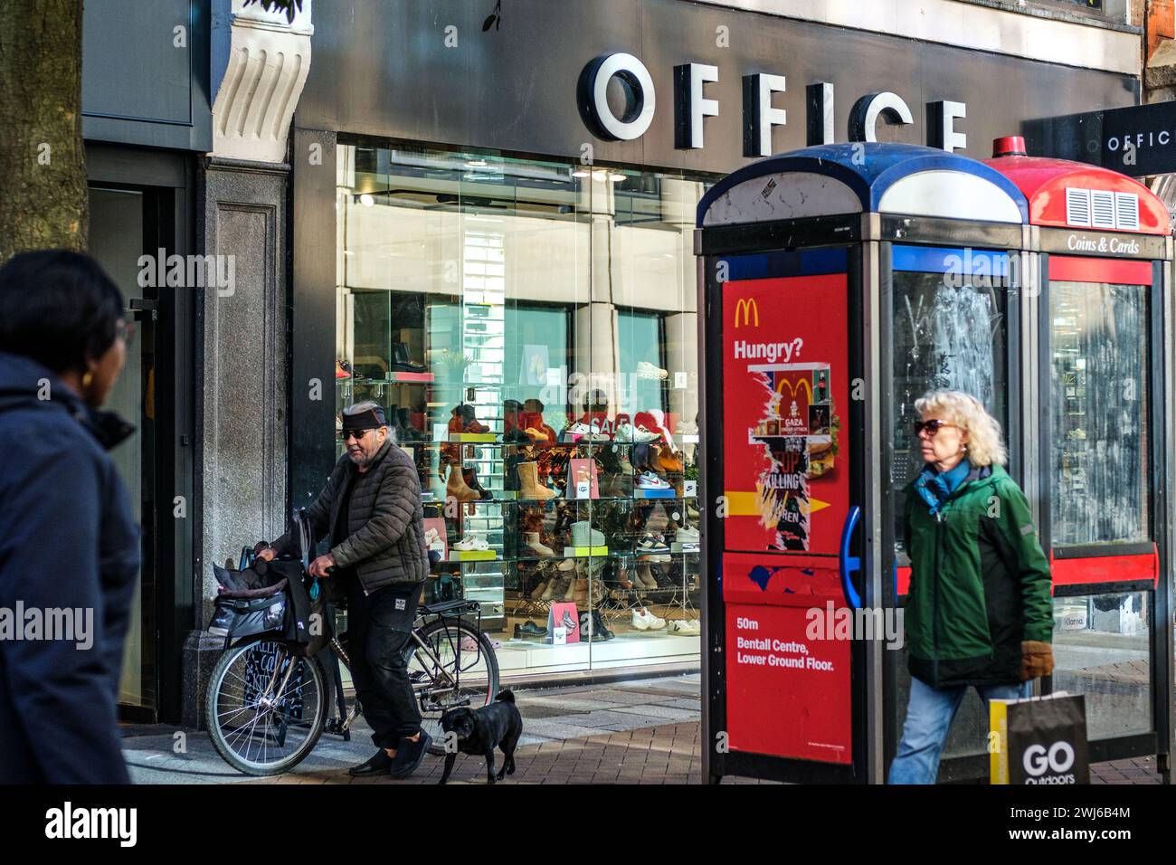 Kingston-upon-Thames, Londres, Royaume-Uni, 12 février 2024, femme portant un sac à provisions et homme sur vélo passant des boîtes téléphoniques publiques et un magasin de chaussures de bureau Banque D'Images