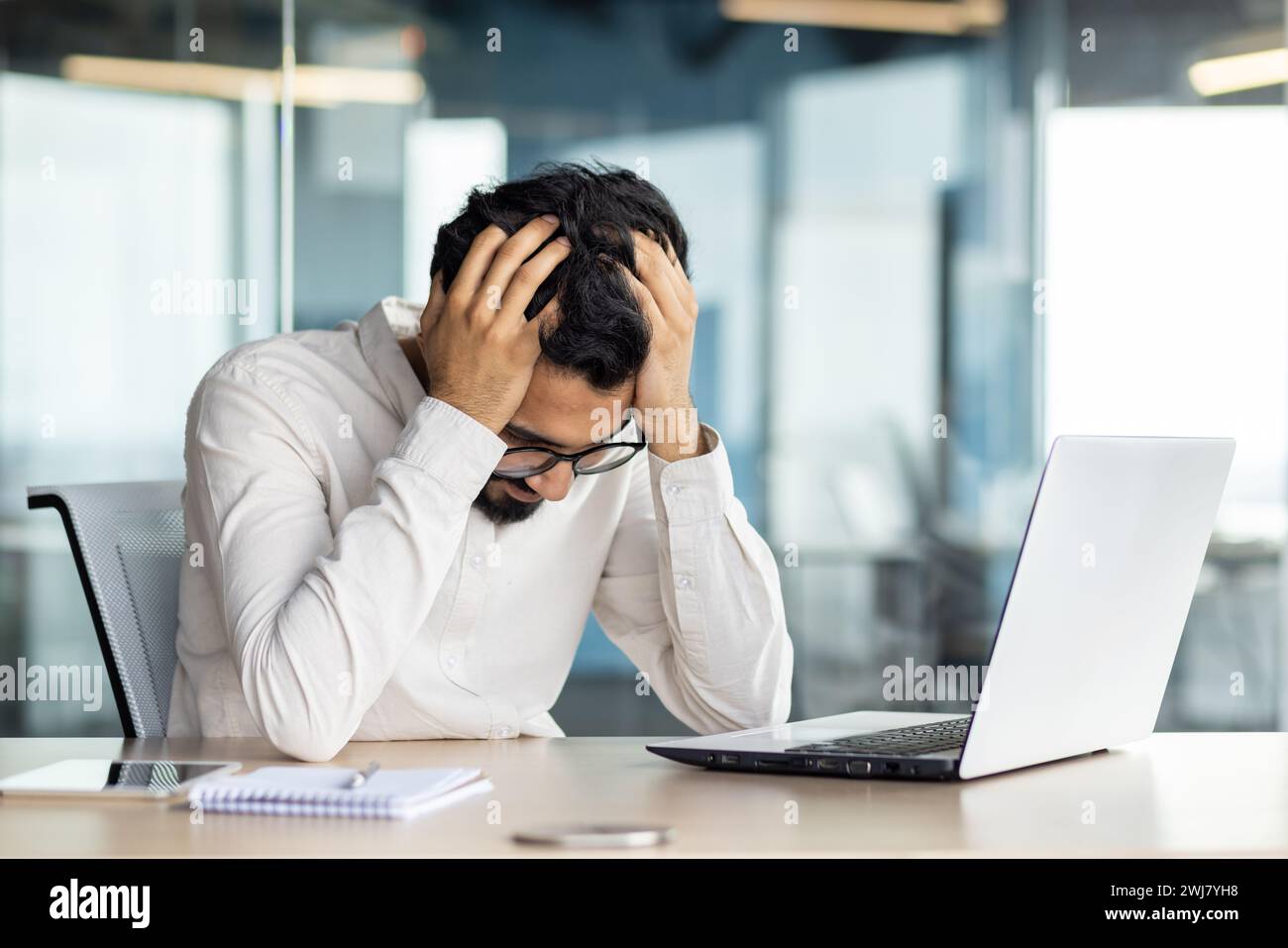 Frustré et inquiet jeune homme d'affaires indien assis au bureau avec ordinateur portable et les mains sur la tête, luttant pour résoudre le problème, fatigué. Banque D'Images