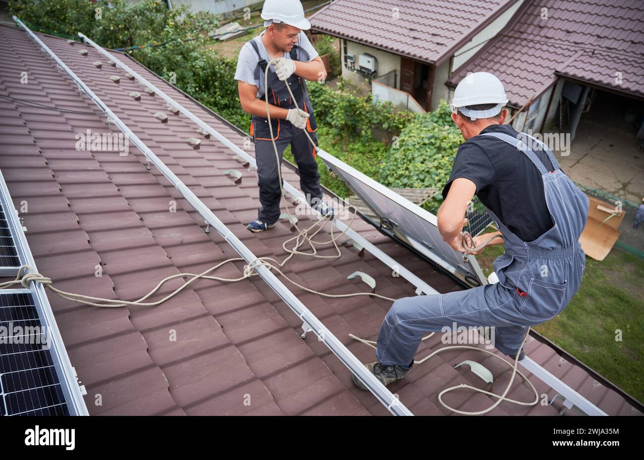 Hommes installateurs soulevant des modules solaires photovoltaïques sur le toit de la maison. Électriciens dans les casques installant le système de panneau solaire à l'extérieur. Concept d'énergie alternative et renouvelable. Banque D'Images
