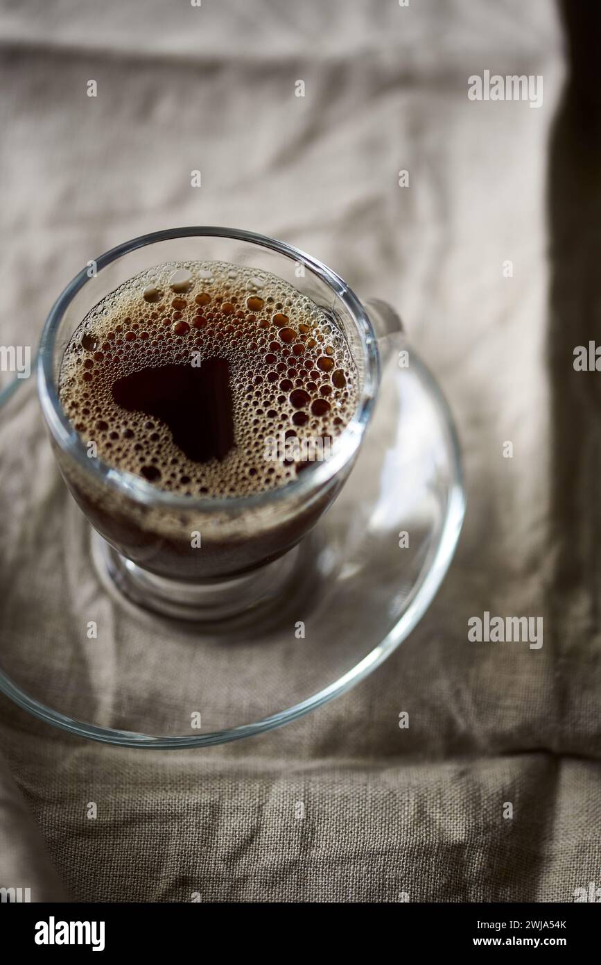 Gros plan détaillé capturant la texture mousseuse d'un café fraîchement préparé dans une tasse en verre transparent sur fond de tissu texturé Banque D'Images