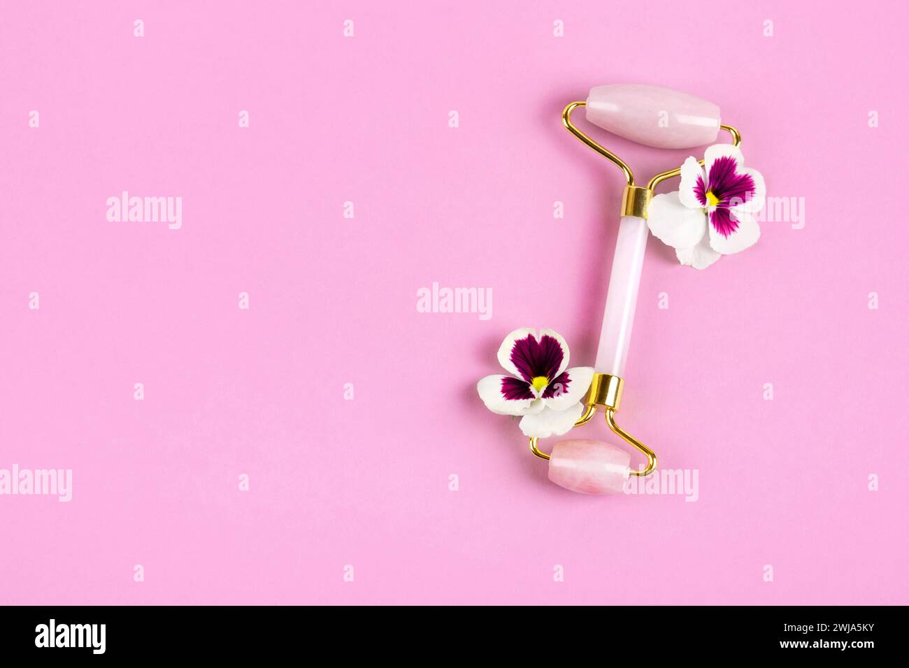 Rouleau de jade rose pour le massage du visage avec des pansies de fleurs. Outils de massage facial. Soin anti âge, lifting et tonifiant. Concept moderne d'autosoins. Banque D'Images