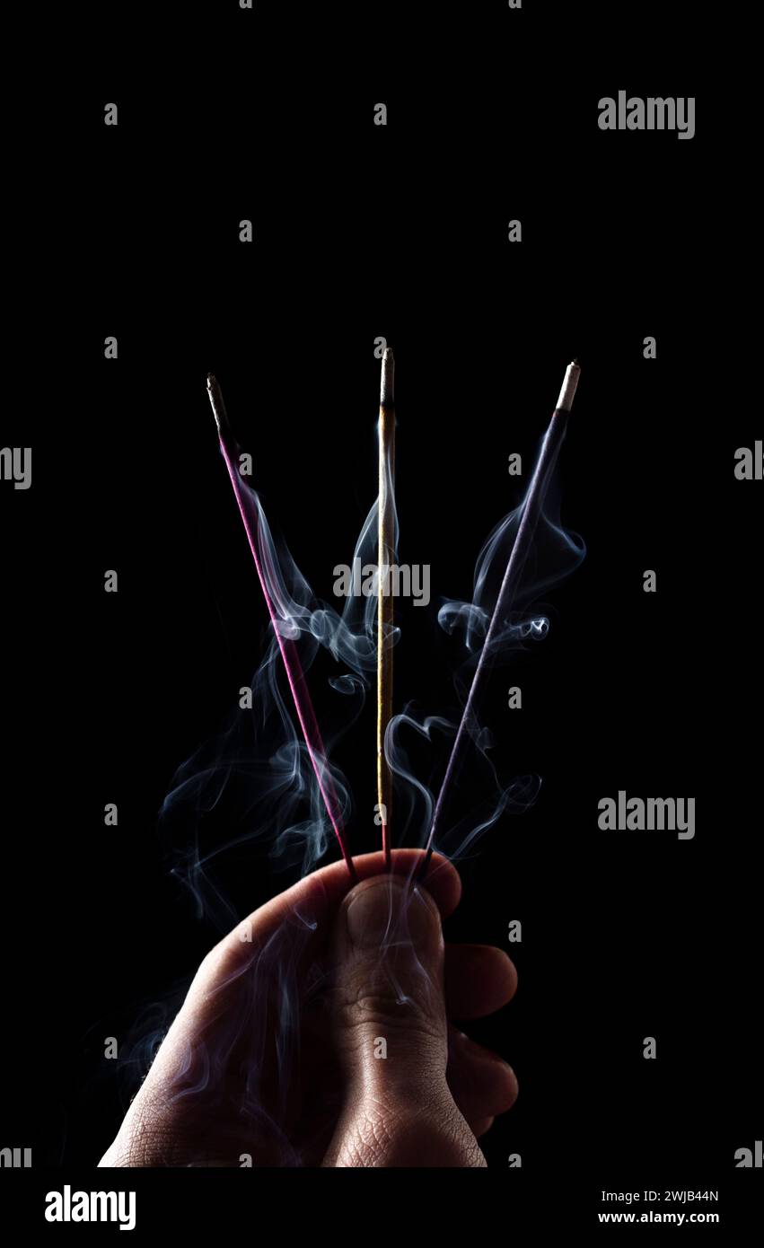 Trois bâtons d'encens multicolores dans la main d'un homme. Les bâtons fument sur un fond noir. Banque D'Images