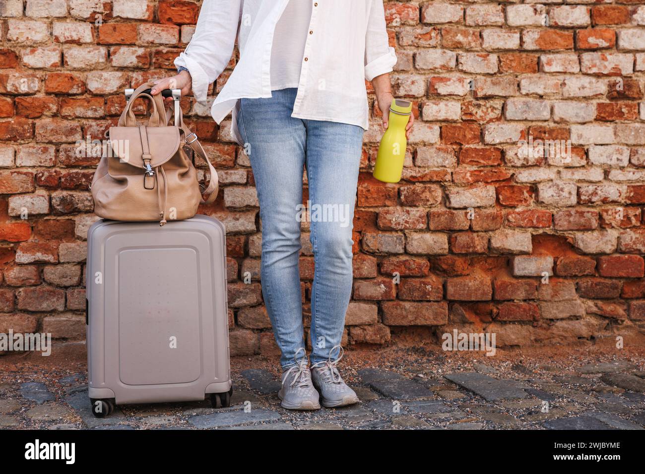 Femme touriste avec valise, sac à dos et bouteille d'eau debout au mur de briques sur la rue de la ville. Concept de voyage et de vacances Banque D'Images