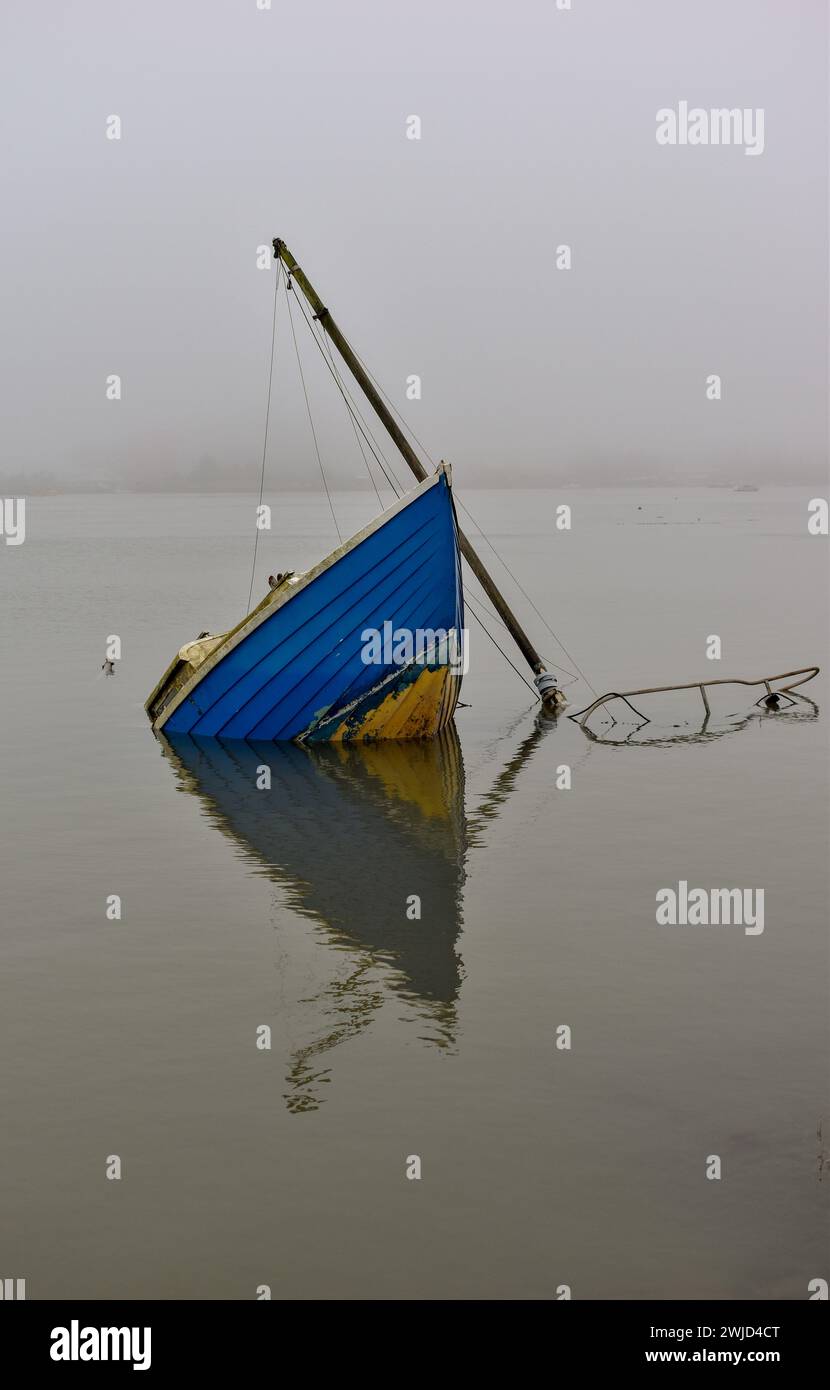 Un petit bateau bleu partiellement submergé par une journée brumeuse avec son reflet visible à la surface de l'eau. Banque D'Images