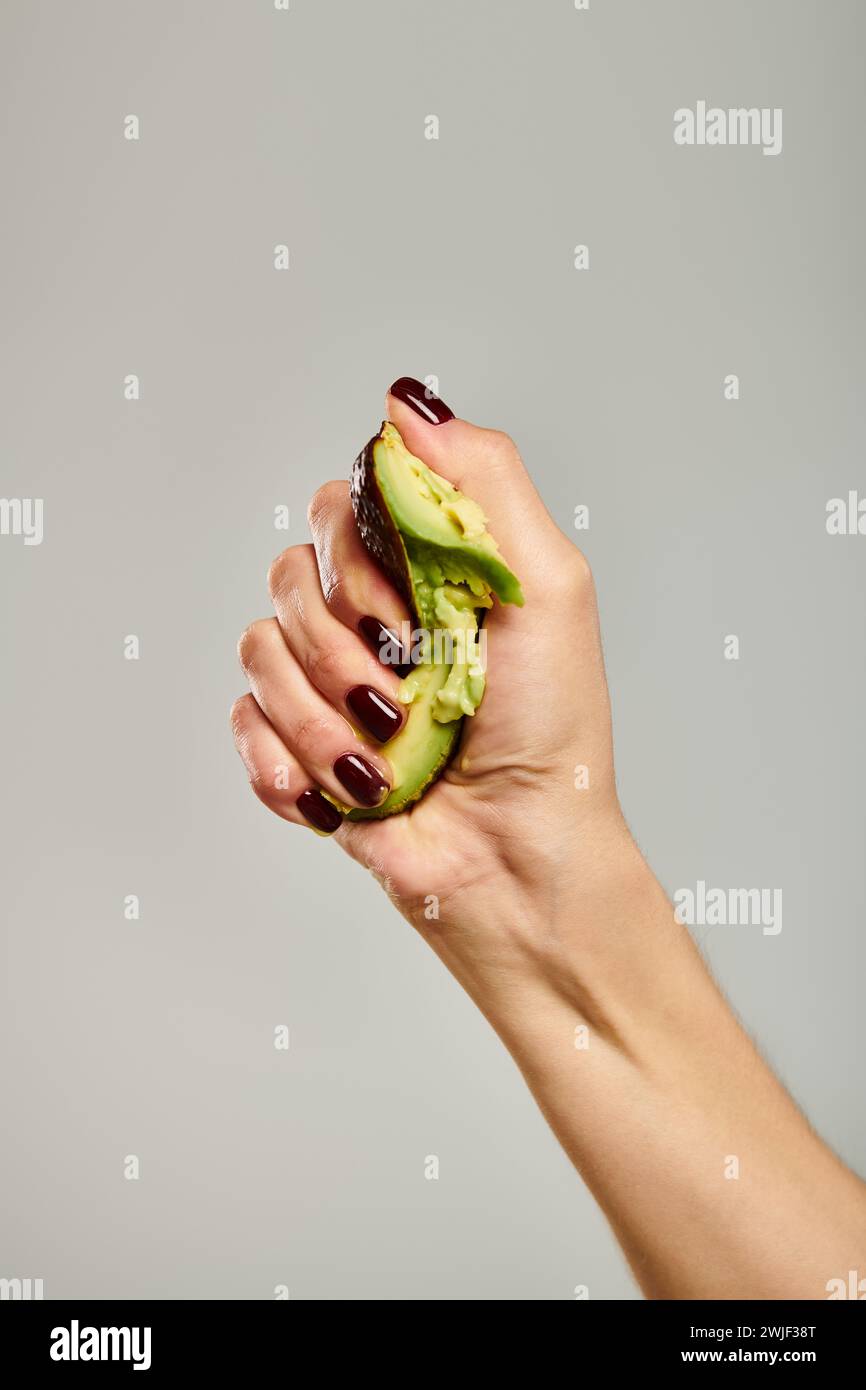 jeune femme inconnue avec du vernis à ongles pressant un avocat vert activement frais sur fond gris Banque D'Images