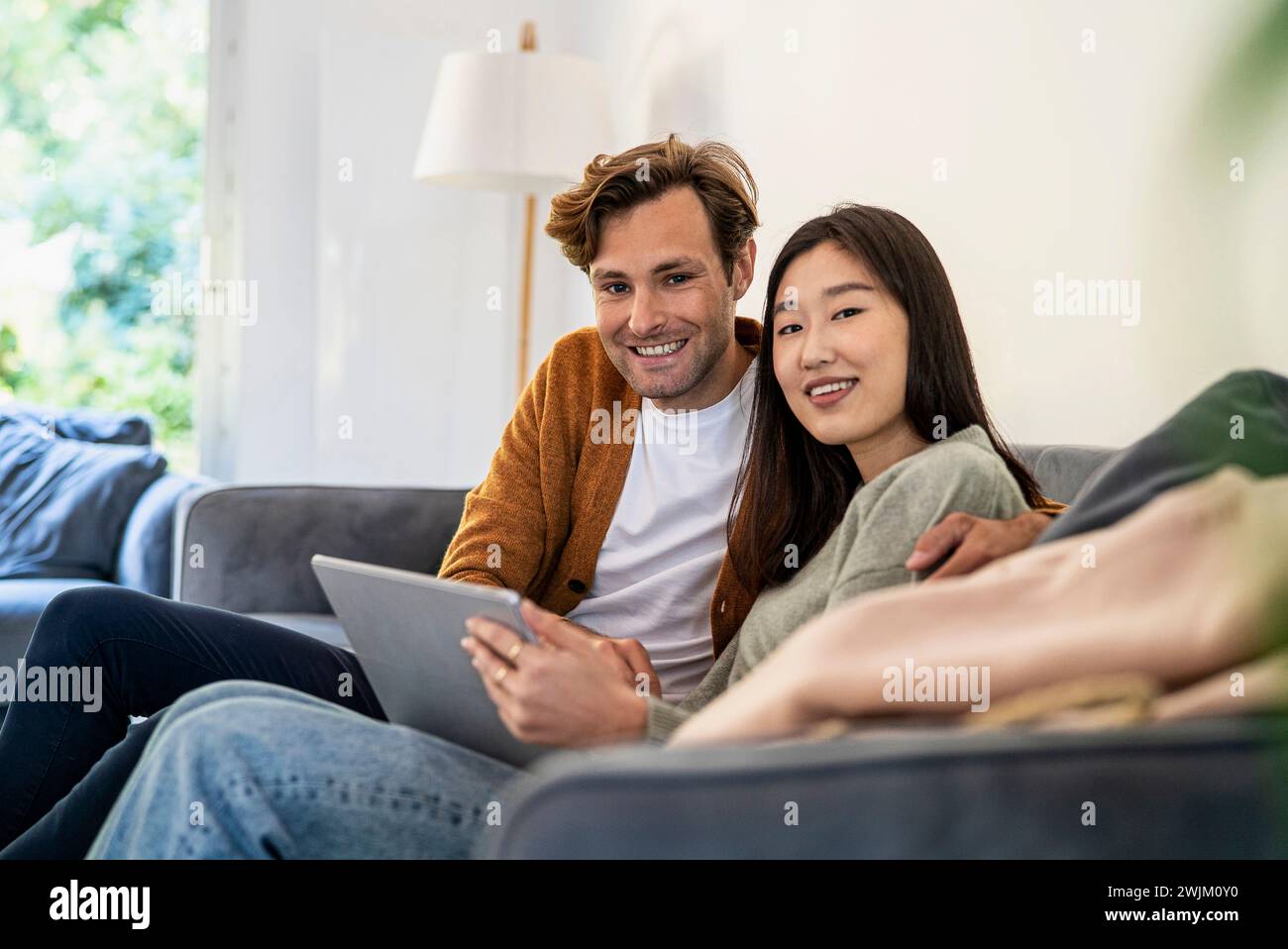 Couple adulte regardant la caméra tout en étant assis sur le canapé à l'aide d'une tablette numérique Banque D'Images