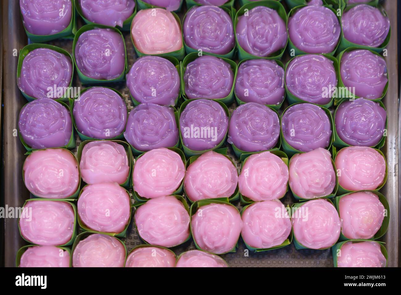 Desserts de gâteau à couche vapeur confectionnés en formes de fleurs, présentés dans des tasses de feuilles de banane, affichant un éventail de couleurs roses et violettes Banque D'Images