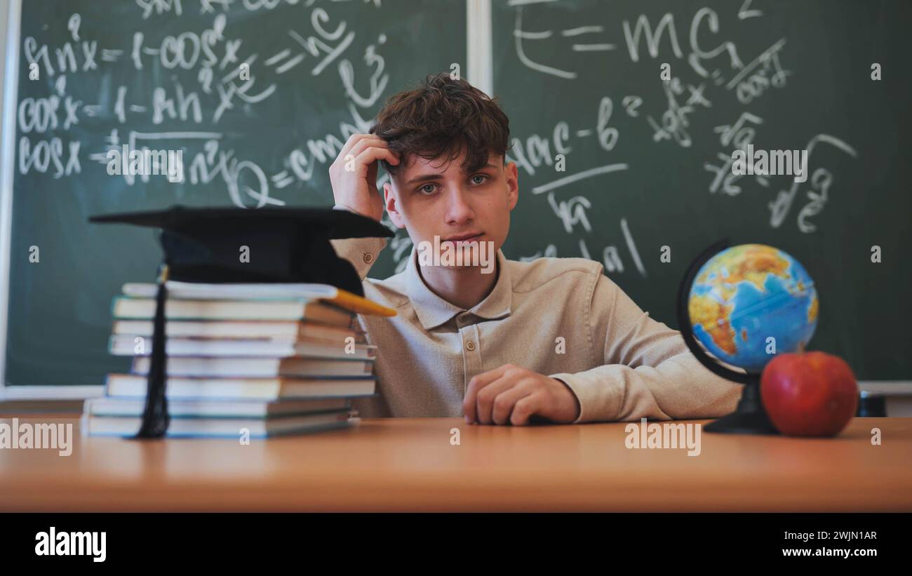 Portrait d'un lycéen sur fond de tableau noir, globe et livres avec casquette. Banque D'Images