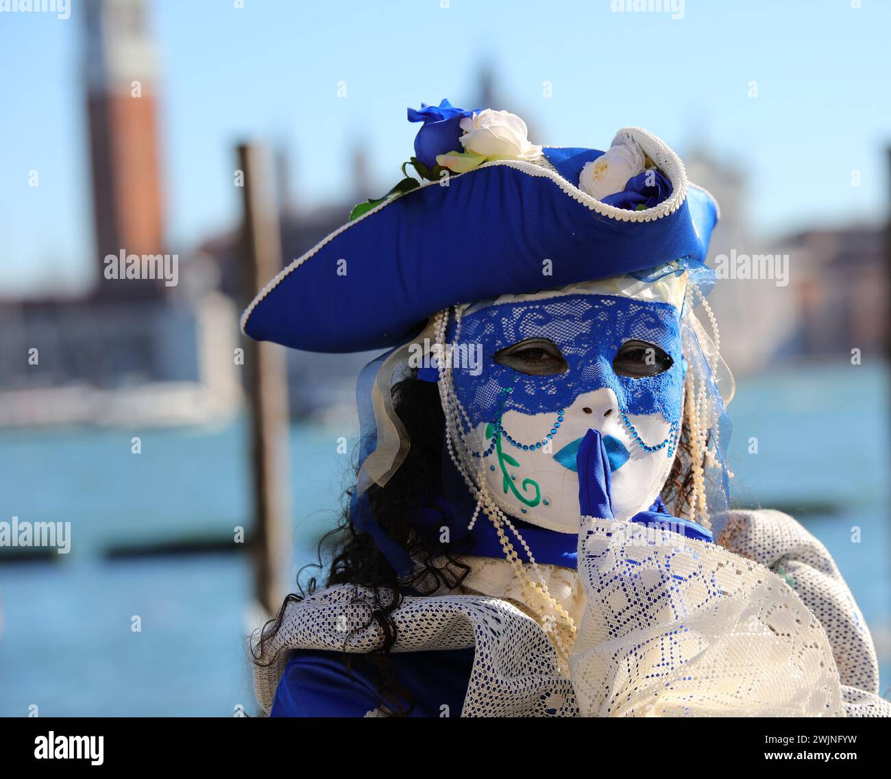 Venise, VE, Italie - 13 février 2024 : femme en masque et chapeau bleu avec le doigt sur les lèvres demandant le silence au bord de la mer Banque D'Images