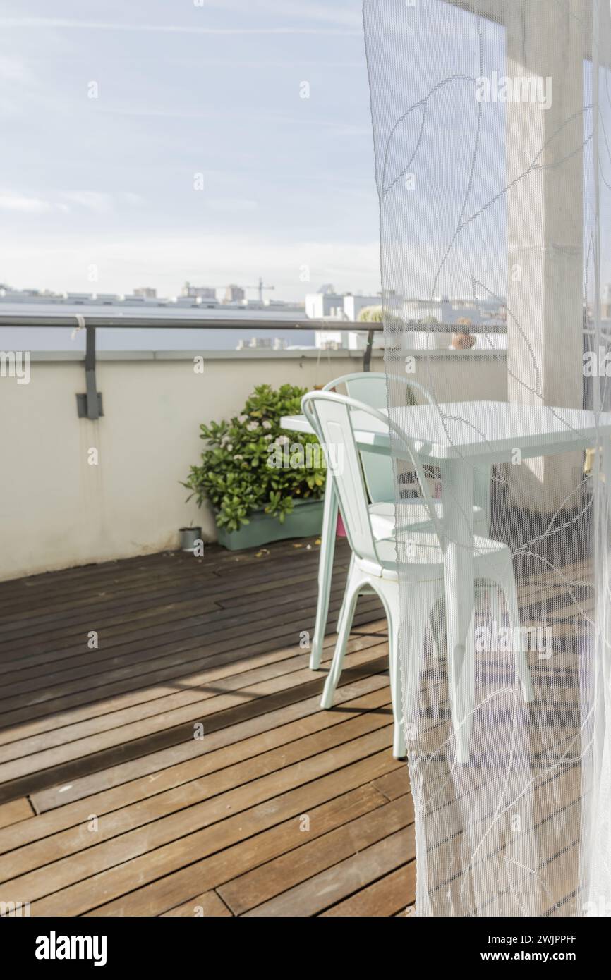 Une jolie table de jardin en métal bleu pastel avec des chaises assorties sur une terrasse urbaine avec parquet, plantes vertes et rideaux translucides Banque D'Images