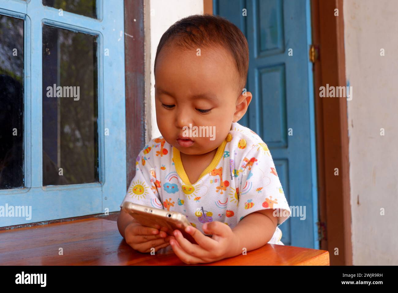 Un garçon de 2 ans joue un smartphone sur une table sur la terrasse d'une maison. Cette photo a été prise à Wadaslintang, Wonosobo, Indonésie. Banque D'Images