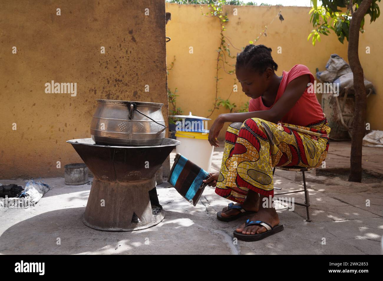 Jeune fille africaine assise devant un simple poêle à charbon de bois attisant le feu ; préparation traditionnelle des aliments Banque D'Images