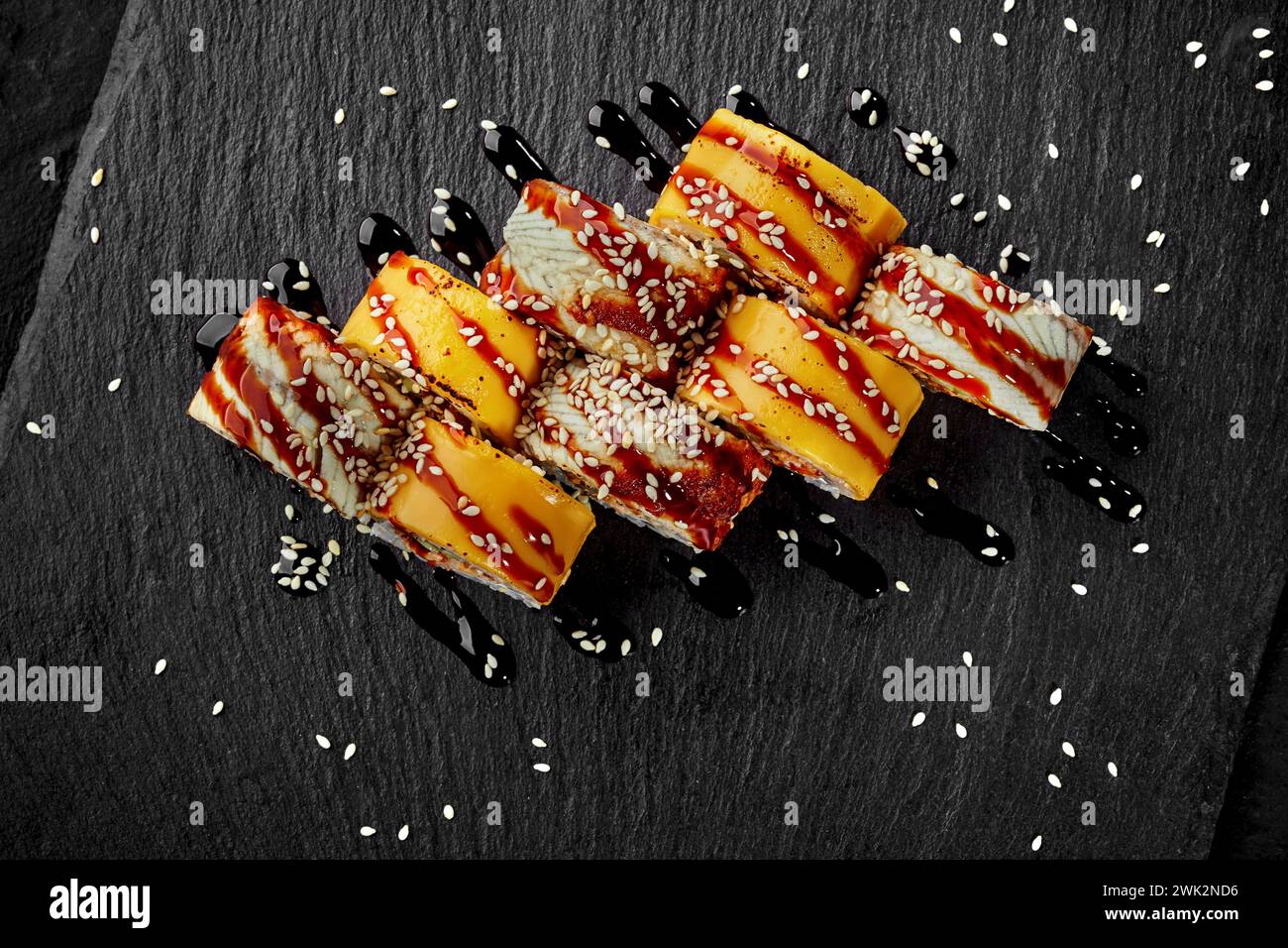 Vue de dessus de rouleaux d'uramaki enveloppés dans des tranches de cheddar et des filets d'anguille habillés de sauce unagi acidulée et saupoudrés de graines de sésame sur le sanglier ardoise noir Banque D'Images