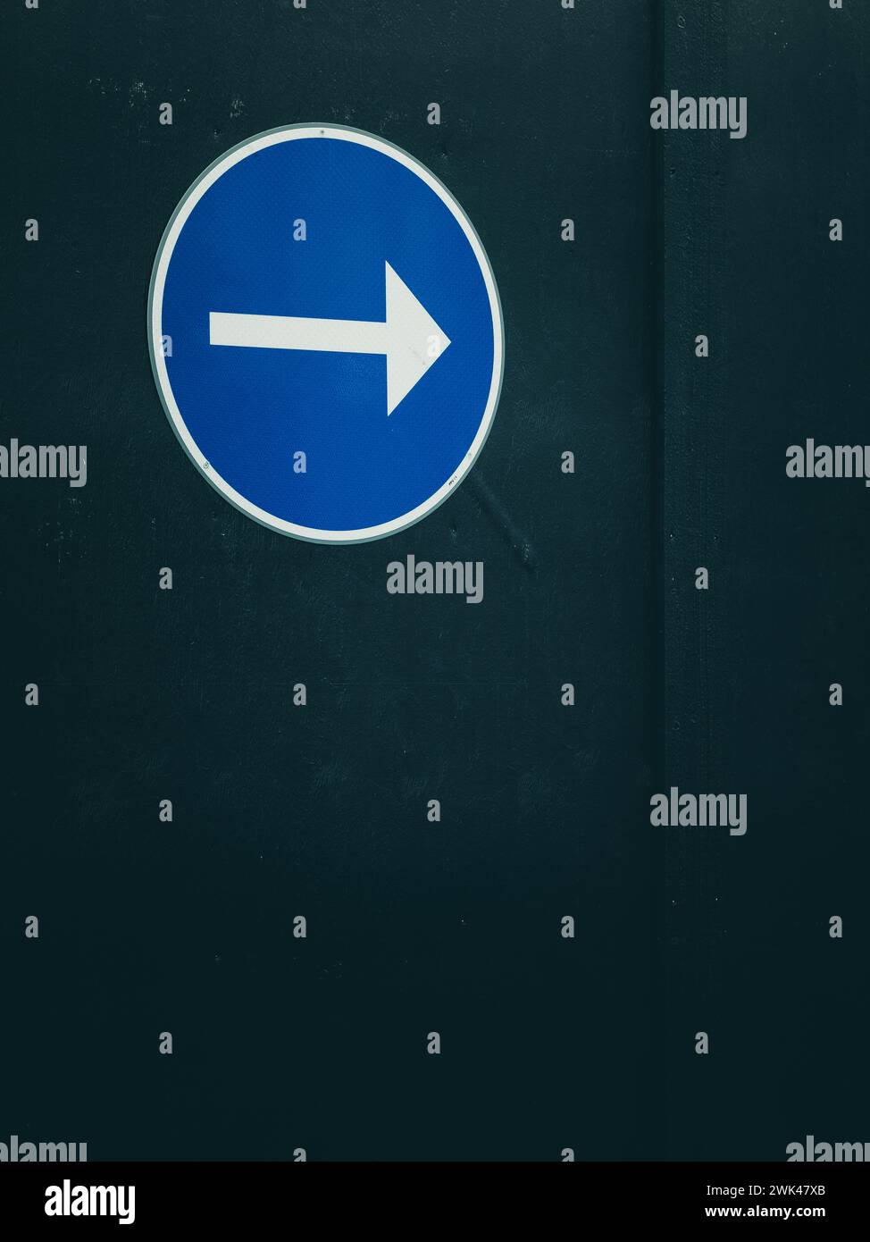 Un signe circulaire bleu proéminent avec une flèche blanche pointant vers la droite est centré dans l'image, apposé sur une surface noire qui remplit l'arrière-plan Banque D'Images