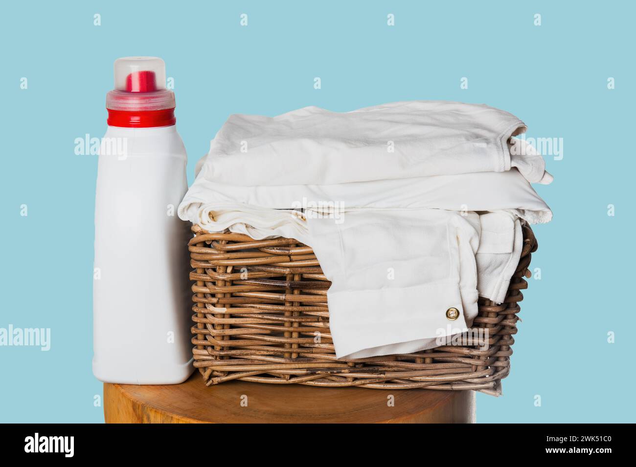 Une bouteille de détergent à lessive à côté d'une pile de vêtements blancs fraîchement lavés dans un panier en osier à fond bleu, un chiffon propre et plié Banque D'Images