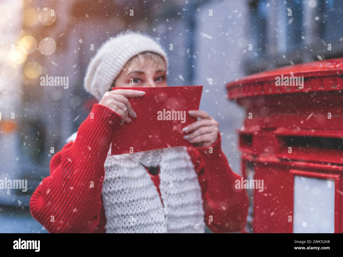 Femme joyeuse dans un manteau rouge et un chapeau blanc mettant une carte dans la boîte aux lettres rouge et se promenant dans une ville anglaise un jour de neige Banque D'Images