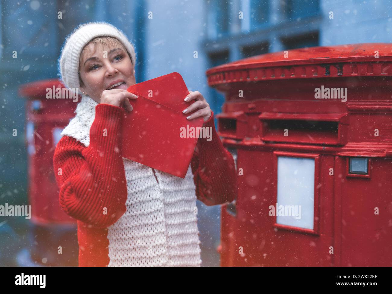 Femme dans un manteau rouge et chapeau blanc mettant une carte dans la boîte aux lettres rouge et se promenant dans une ville anglaise un jour de neige Banque D'Images