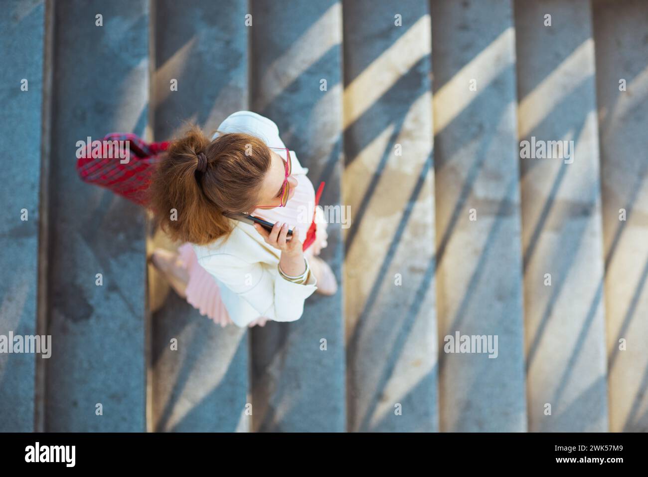 Vue supérieure de l'élégante femme d'âge moyen en robe rose et veste blanche dans la ville avec sac rouge et lunettes de soleil parlant sur un smartphone. Banque D'Images