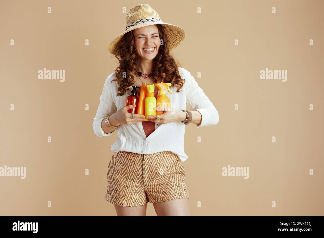 Vacances à la plage. femme élégante heureuse en blouse blanche et short isolé sur fond beige avec crème solaire et chapeau de paille. Banque D'Images