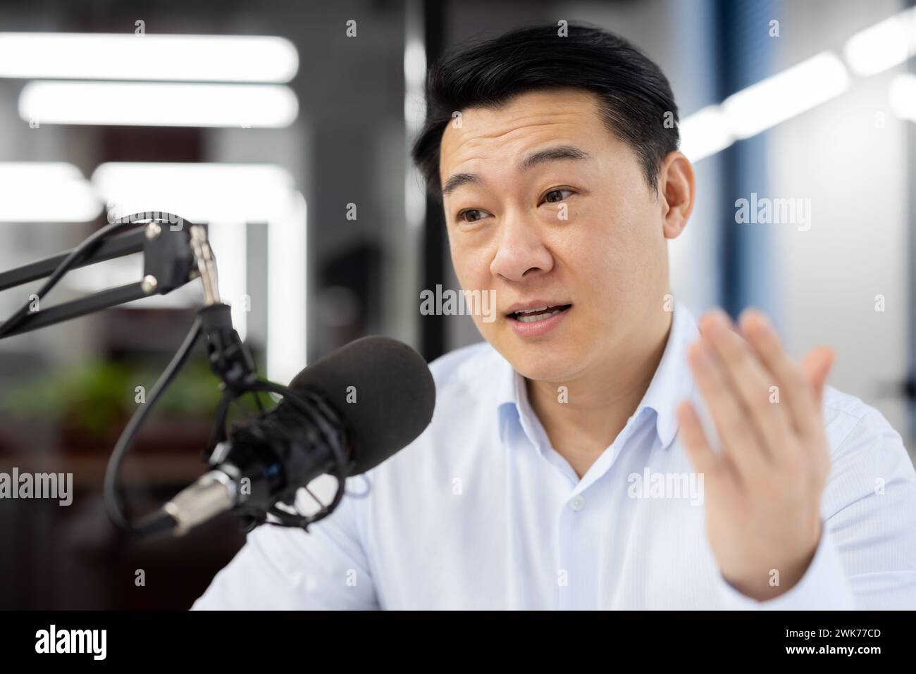 Photo en gros plan d'un jeune homme asiatique assis dans le bureau à un bureau devant un micro et ayant une réunion en ligne par appel vidéo, discutant, expliquant, faisant des gestes avec sa main. Banque D'Images