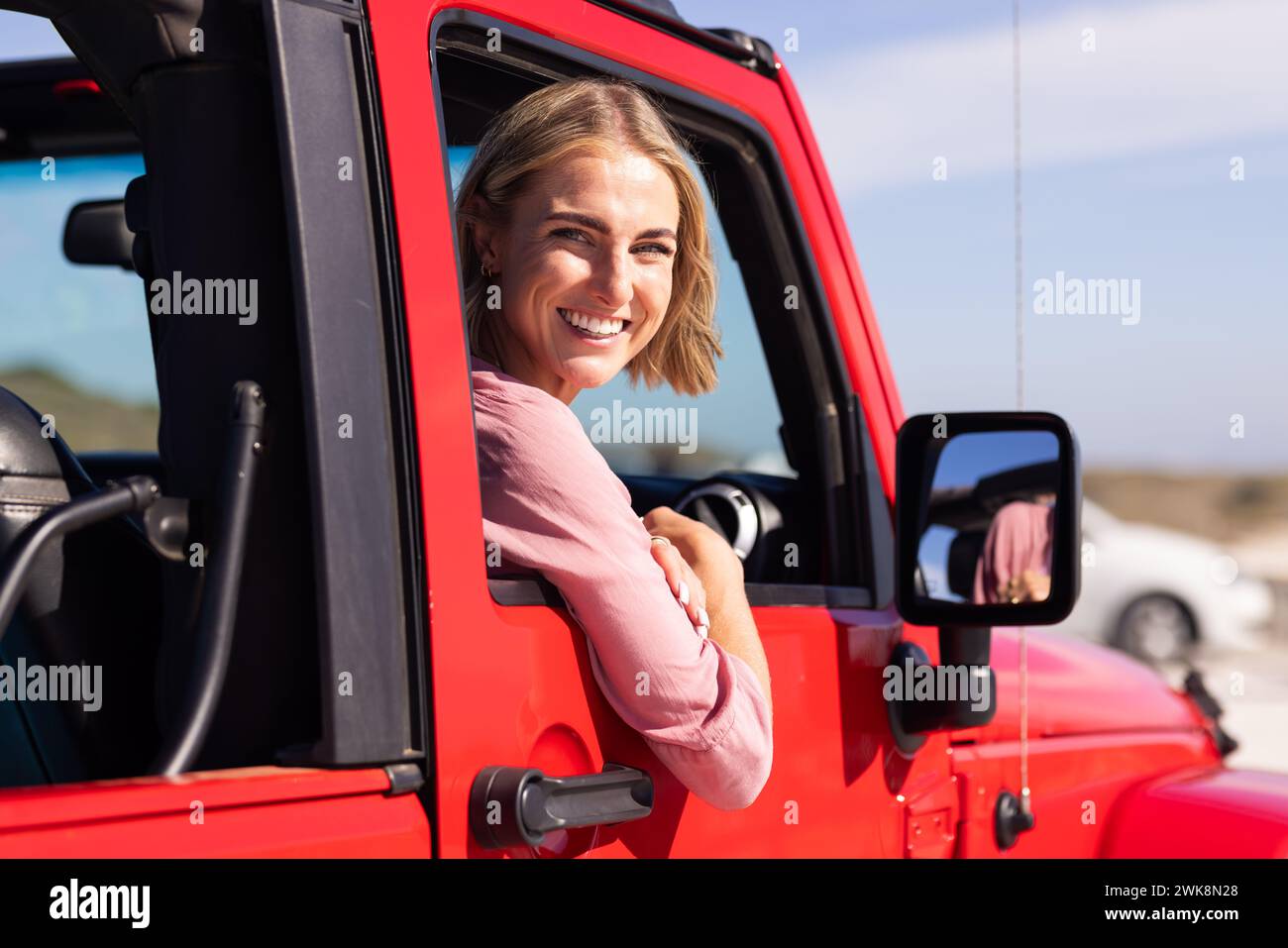 Une jeune femme caucasienne sourit depuis le siège du conducteur d'un véhicule rouge lors d'un voyage sur la route Banque D'Images