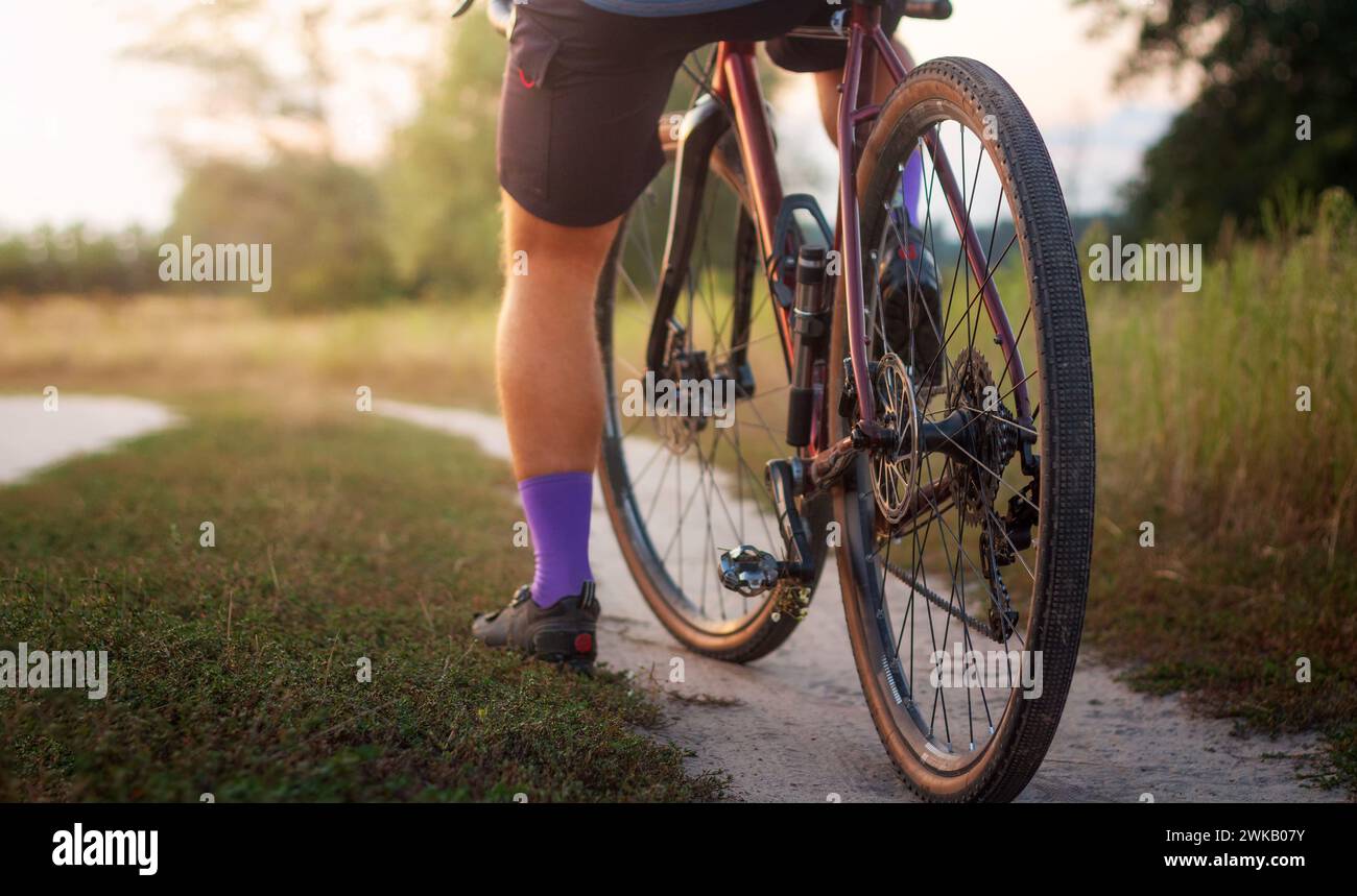 Cycliste professionnel sur son vélo debout sur un chemin de terre dans un champ au coucher du soleil. Sport et mode de vie actif. Banque D'Images