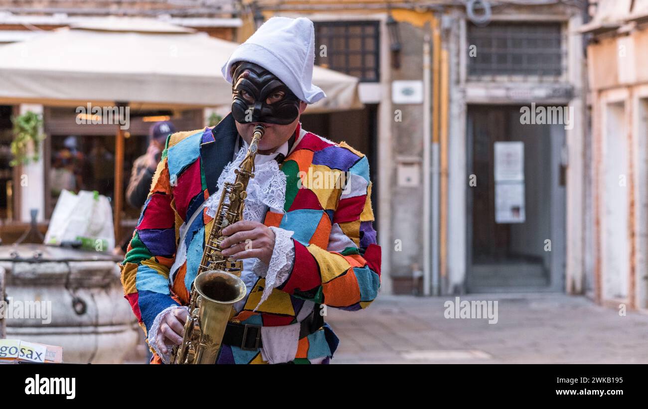 Journée ensoleillée au Carnaval de Venise avec de beaux masques et un artiste de rue habillé en Arlequin jouant du sax. ArlecchinoDiagoSax. Banque D'Images