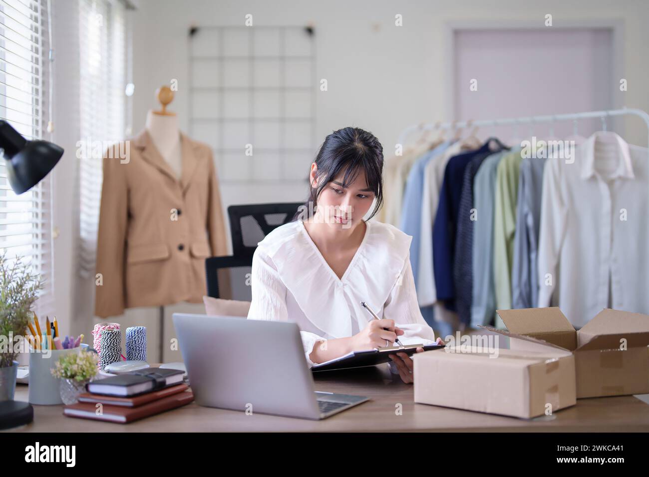 Jeunes femmes marchandes asiatiques vérifiant la commande d'achat en ligne du client sur ordinateur portable et écrivant des informations dans le presse-papiers tout en travaillant et en expédiant la livraison Banque D'Images