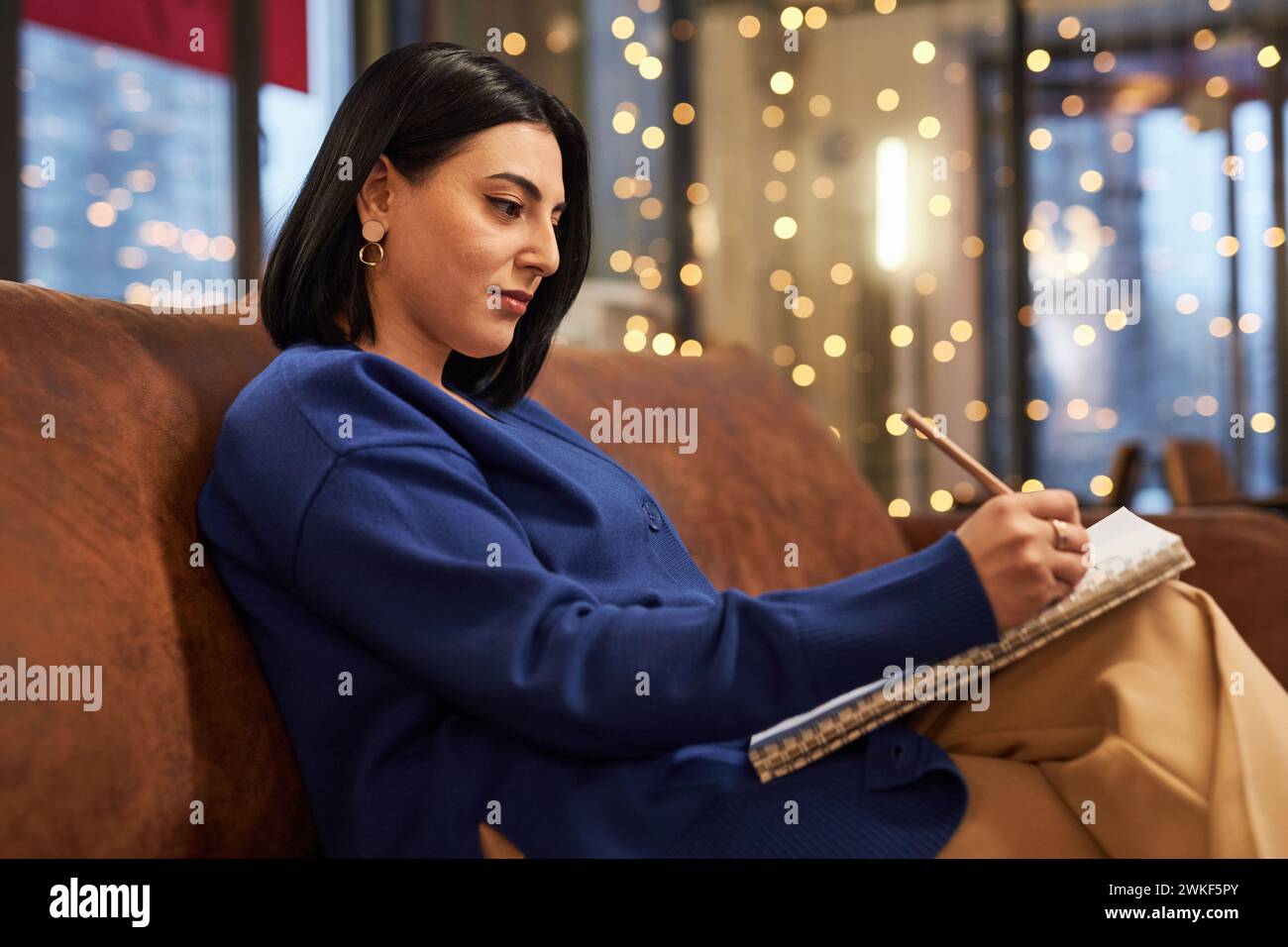Portrait de vue de côté de la jeune femme du moyen-Orient écrivant dans un cahier ou dessinant un croquis tout en étant assise sur un canapé dans un café Banque D'Images