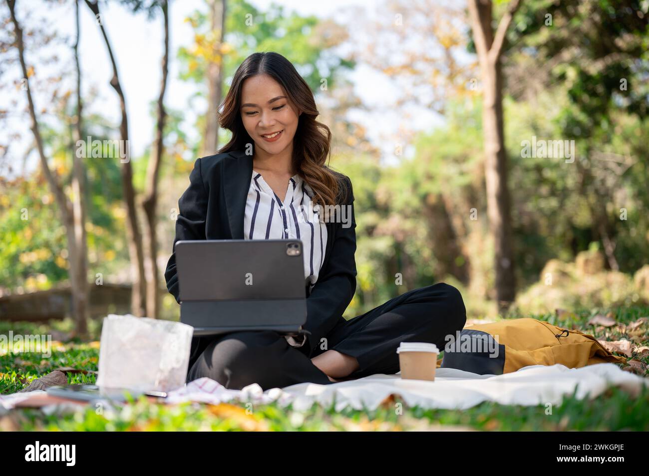 Une femme d'affaires asiatique positive et attrayante travaillant à distance dans un parc, travaillant sur sa tablette numérique sur un tapis de pique-nique. Banque D'Images