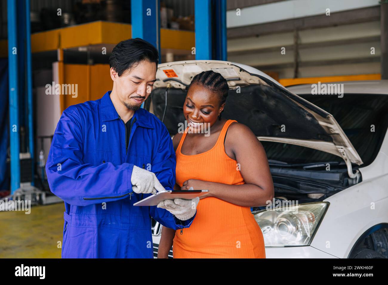 Les femmes clientes noires multiethniques heureuses apprécient avec la liste de contrôle de service de voiture de travailleur de mécanicien asiatique dans l'atelier de centre automatique Banque D'Images