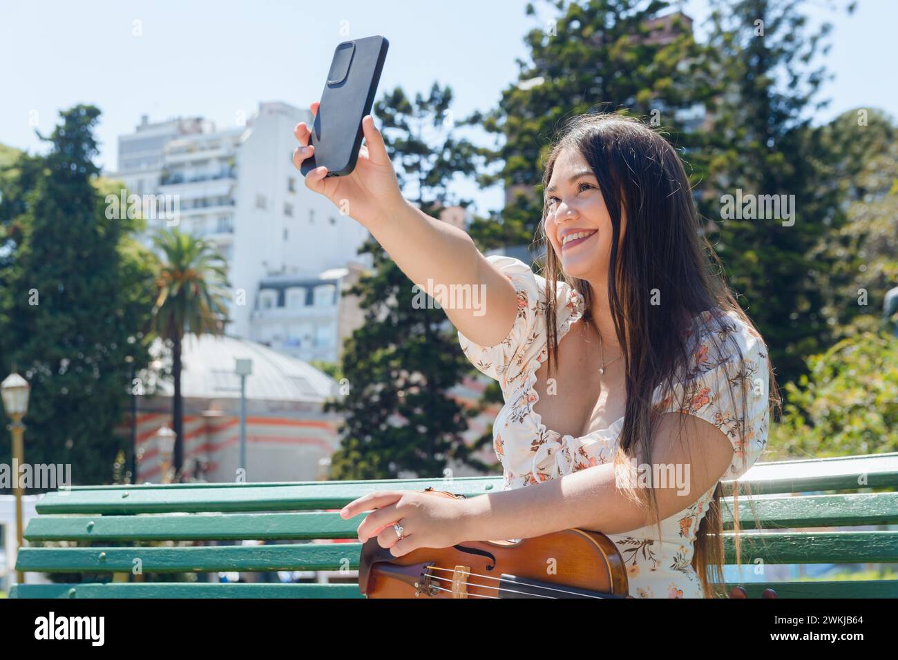 Jeune femme violoniste latina busker assise avec son violon sur un banc à l'extérieur, sur la plaza, souriant, prenant une photo selfie avec son téléphone, la technologie conce Banque D'Images