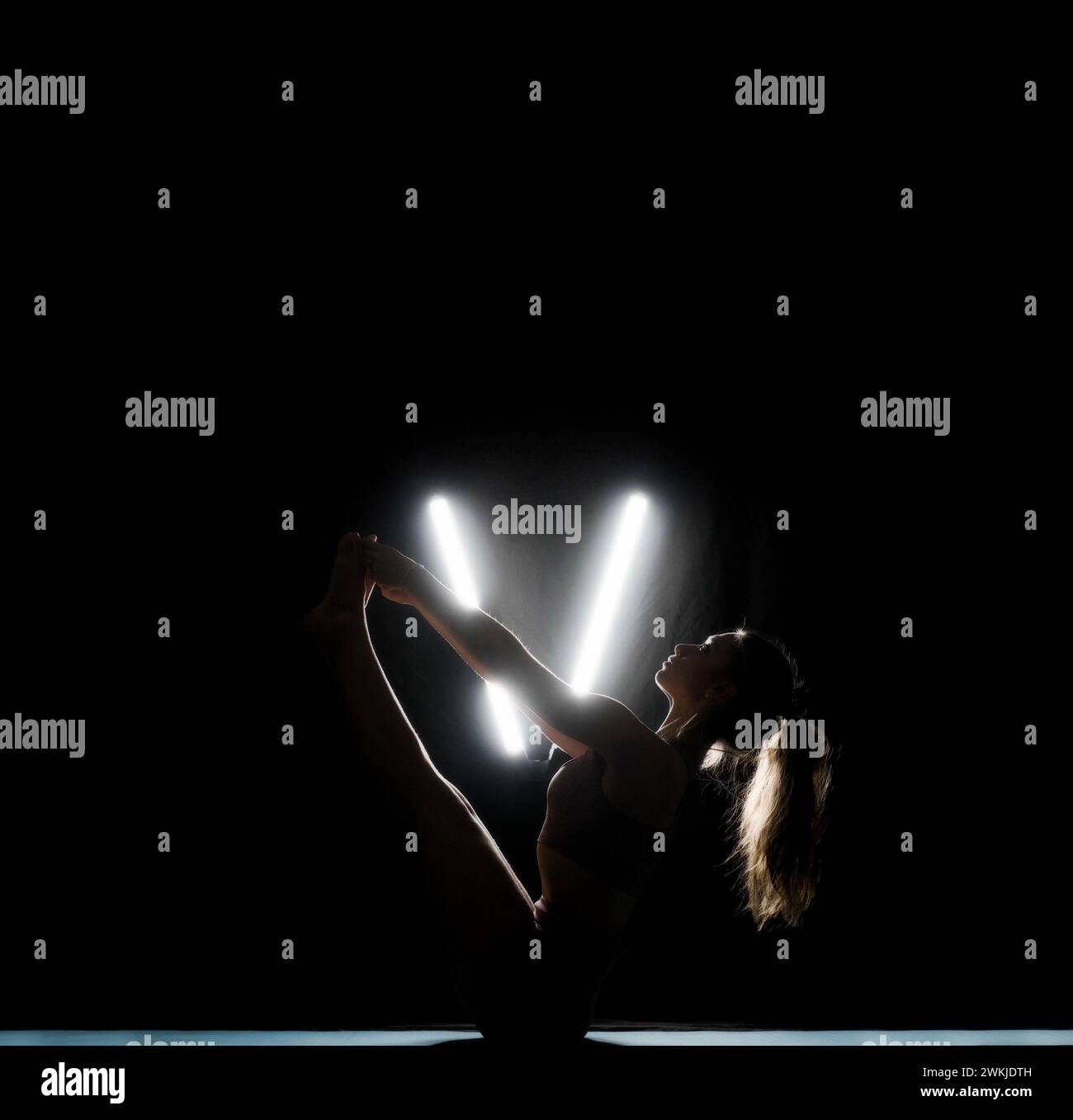 Une femme silhouette étire ses jambes dans une pose de yoga sur un fond sombre éclairé par des lumières blanches en forme de V néon. Banque D'Images