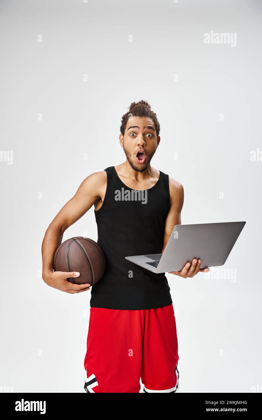 sportif afro-américain surpris tenant basket-ball et ordinateur portable regardant la caméra, paris en ligne Banque D'Images